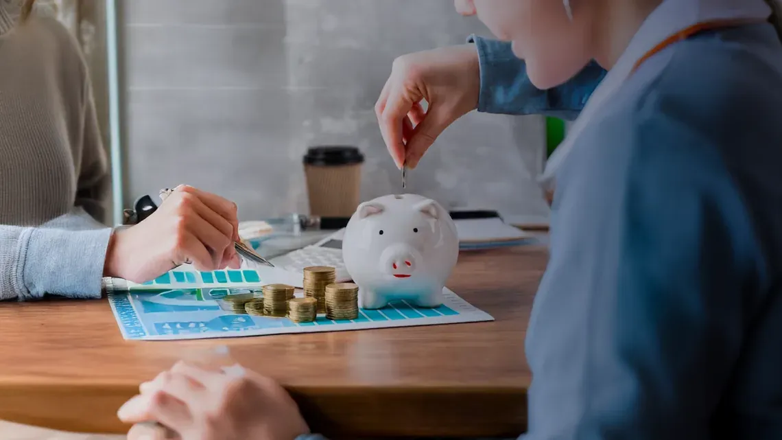 Persona deposita una moneda en una alcancía sobre un escritorio con monedas y gráficas.